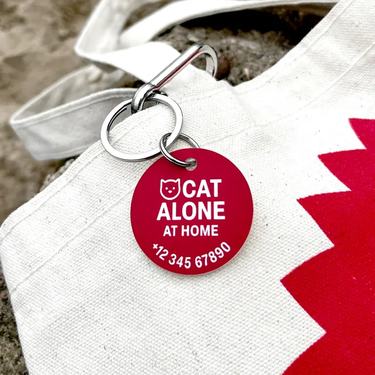 Roter personalisierter Notfallkontaktanhänger für Katze mit weißer Gravur ‚Cat alone at home‘, befestigt an Jutebeutel auf Boden, silberner Metallkarabiner
