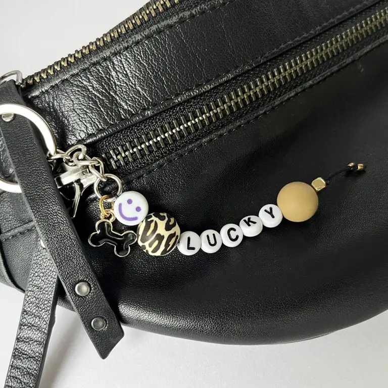 Personalisierter Perlen-Schlüsselanhänger mit Hundenamen, schwarzem Knochen-Charm, Leoparden-Look-Perle, ockerfarbener Silikonperle, Smiley-Perle und Edelstahlring, hängend an einer Leder-Crossbody-Bag